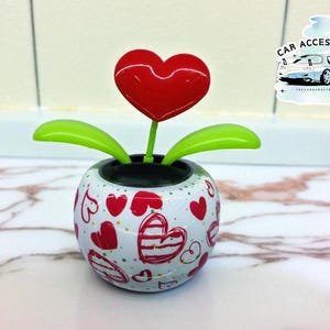 May include: A decorative solar-powered car accessory shaped like a flower with a red heart-shaped top and green leaves. The white base is decorated with red hearts. The image also includes a sticker with the text "CAR ACCESSORIES".