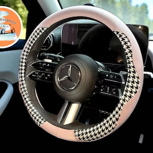 May include: A car steering wheel with a light pink cover and a black and white houndstooth pattern. The steering wheel has a black center with the Mercedes-Benz logo. The car interior is visible in the background.