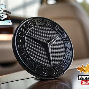 May include: Black Mercedes-Benz emblem with a textured surface. The emblem is round and has the words "Mercedes-Benz" written around the edge.