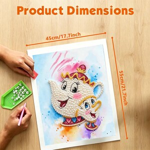 K&ouml;nnte beinhalten: Ein Diamond-Painting-Set mit einem Teekannen- und Tassenmotiv aus wei&szlig;en perlenartigen Steinen, das 45 cm mal 55 cm misst. Das Set enth&auml;lt eine gr&uuml;ne Ablageschale mit Steinen, einen rosa Applikatorstift und eine bedruckte Leinwand.