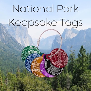May include: A collection of colorful National Park Keepsake Tags on a silver wire ring. Each tag features a park name, date, and illustration, including Yosemite, Zion, and Badlands. The background shows a scenic mountain view.