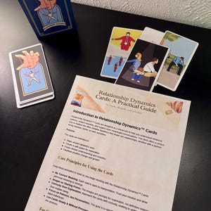 May include: A deck of Relationship Dynamics Cards with a navy blue box featuring a graphic of a hand controlling a person. Several cards are displayed, each with unique illustrations. A guide titled "Relationship Dynamics Cards: A Practical Guide" is also present.