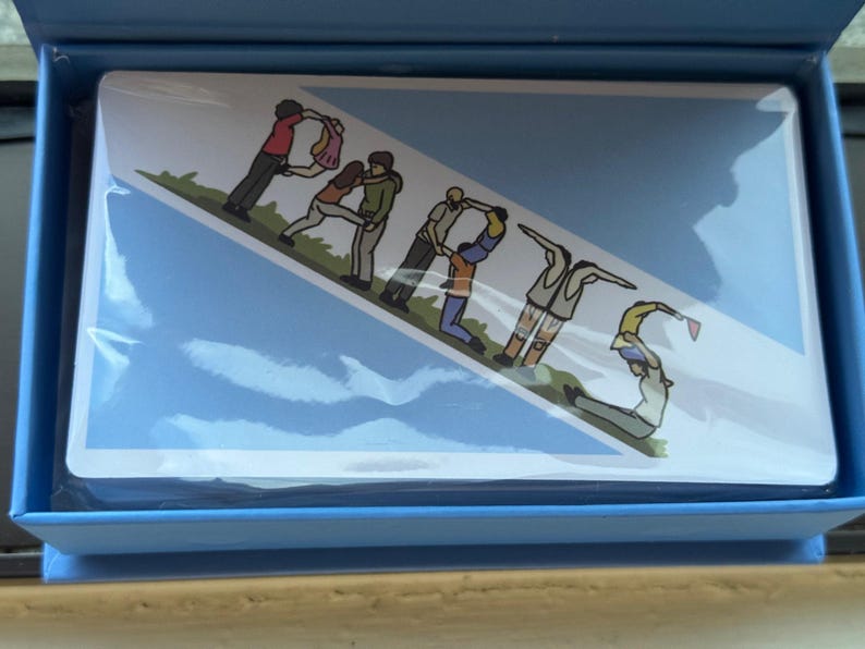 May include: A blue card with a white border and a graphic of people forming the word "PARIS" in a line. The people are wearing different colored clothing and are standing on a green grassy area. The background is a light blue sky with white clouds.