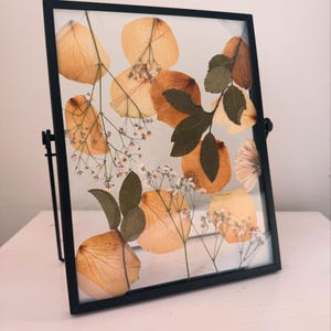 Op de afbeelding: Een zwart ingelijst beeld met een gedroogde bloemenarrangement. De bloemen zijn voornamelijk in tinten geel en bruin, met enkele kleine witte bloemen. Het frame heeft een standaard aan de achterkant.