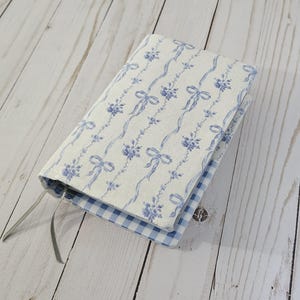 May include: A fabric book cover with a blue and white floral pattern and a blue gingham interior. The cover features a repeating design of blue bows and flowers on a white background. A gray ribbon bookmark is attached.