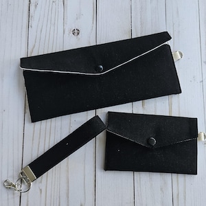 Könnte beinhalten: Ein Set aus drei schwarzen Stoffaccessoires: eine lange Geldbörse, ein kleiner Kartenhalter und eine Handschlaufe mit silberfarbenem Verschluss. Die Geldbörse und der Kartenhalter haben einen weißen Rand und einen Knopfverschluss.