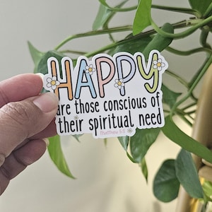May include: A sticker with the word "HAPPY" in colourful letters, decorated with small daisy flowers. The text "are those conscious of their spiritual need" is below, with "Matthew 5:3" at the bottom. The sticker is held in front of green leaves.