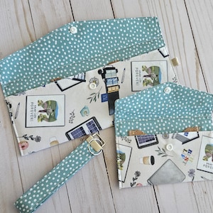 Puede incluir: Un conjunto de tres accesorios de tela: una cartera grande, una cartera pequeña y una correa de muñeca. Los artículos presentan un fondo blanco con un estampado de sellos, bolígrafos y ordenadores portátiles. El interior y la correa son de color turquesa con lunares blancos.