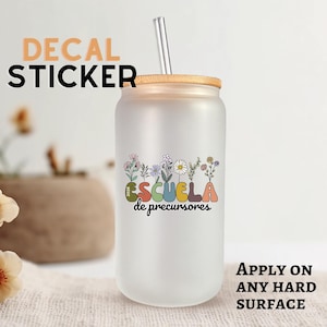 May include: Frosted glass tumbler with a bamboo lid and a metal straw. The tumbler features a floral decal with the text "ESCUELA de precursores". The words "DECAL STICKER" and "APPLY ON ANY HARD SURFACE" are also visible.