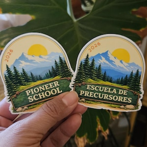 May include: Two stickers with a mountain and forest design. One reads "Pioneer School" and the other "Escuela de Precursores". Both have a yellow sun and the year "2026" printed on them. The stickers are held by a hand.