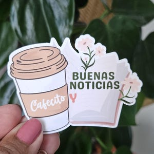 May include: A sticker with a cartoon illustration of a cup of coffee and an open book. The coffee cup has the text "Cafecito" on it. The book has the text "Buenas Noticias y" on it. The sticker has a pink and white floral design.