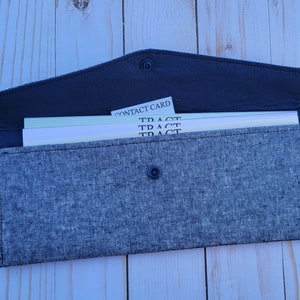 Puede incluir: Una cartera de lino gris con una solapa de cuero negro y un cierre a presión. La cartera tiene un bolsillo con cremallera en el lateral y un cierre de metal. La cartera está abierta y contiene varias tarjetas de visita blancas con el texto "CONTACT CARD" y "TRACT" impreso en ellas.