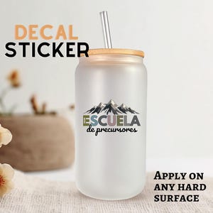 May include: Frosted glass tumbler with a bamboo lid and a metal straw. The tumbler features a mountain graphic with the text "ESCUELA de precursores". The words "DECAL STICKER" are above the tumbler.
