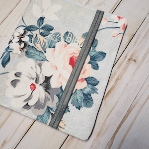 May include: A floral-patterned book cover with the text "LA BIBLIA" in white. The cover features large, light pink and white flowers with green and blue leaves against a light gray background. A gray elastic band secures the cover.