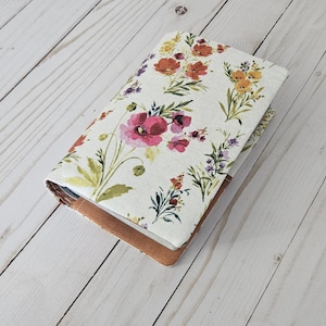 Può includere: Una copertina di diario con motivo floreale, con un design ad acquerello di fiori rossi, rosa, gialli e viola su sfondo bianco. La copertina è chiusa, rivelando un bordo in pelle marrone. Il diario è appoggiato su una superficie di legno chiaro.