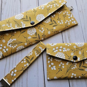 Puede incluir: Un conjunto de tres accesorios a juego: una cartera larga, una cartera pequeña y una correa de muñeca. Todos los artículos presentan un estampado floral amarillo mostaza con flores blancas y detalles en negro. Cada cartera tiene un cierre de botón a presión negro.