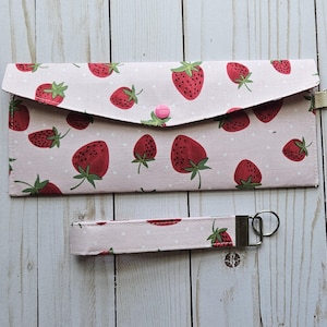 Puede incluir: Un monedero rosa y una pulsera a juego con un estampado de fresas rojas y hojas verdes sobre un fondo rosa claro. El monedero tiene un cierre de solapa con un botón rosa.