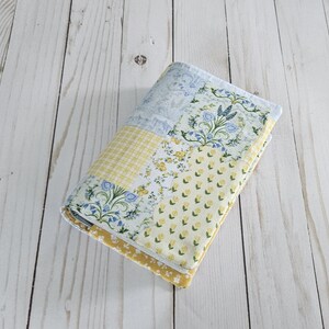 May include: A rectangular fabric book cover featuring a patchwork design of various floral and checkered patterns in shades of yellow, light blue, and white. The quilted textile accessory displays repeating motifs of small flowers and grid lines.