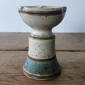 May include: A small, white ceramic pedestal with a brown rim and a blue band around the base. The pedestal is speckled with brown and white flecks.