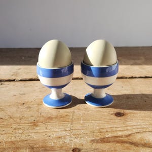 May include: Two ceramic egg cups with blue and white stripes, each holding a pale egg. The egg cups have a pedestal base and are set on a wooden surface.