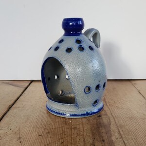 Op de afbeelding: Een keramische kandelaar met een blauw en grijs gespikkelde afwerking. De houder heeft een ronde body met een grote opening en diverse kleinere gaten. Het heeft een handvat en een blauwe bovenkant.