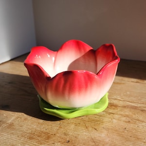 以下が含まれることがあります： 赤と白のグラデーションが施された、花型のセラミックボウル。緑色の葉の形をしたベースの上に置かれています。ボウルは木製の表面にあり、デザインは咲き誇る花に似ています。インテリアデコレーションに最適です。