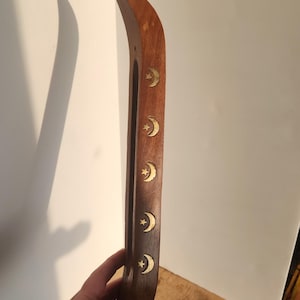May include: A curved, wooden incense holder with inlaid brass crescent moon and star designs. The holder is a rich brown color and is held by a person's hand.