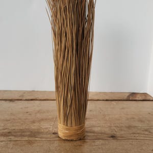 May include: A bundle of dried, thin, light brown reeds, tightly bound at the base with a woven band. The reeds stand upright, creating a cylindrical shape. The background is a plain white wall and a wooden surface.