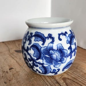 May include: A round, white ceramic vase with a blue floral design. The vase features a wide opening and a bulbous body. The blue floral pattern includes large flowers and swirling leaves. The vase sits on a wooden surface.