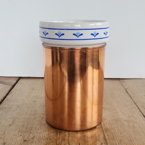 May include: A cylindrical container featuring a polished copper body and a white ceramic rim. The ceramic section is decorated with a horizontal blue line and a repeating pattern of small blue floral motifs encircling the top edge of the vessel.