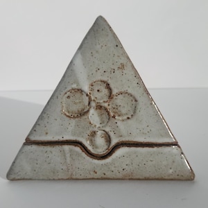 May include: A triangular ceramic incense holder with a white glaze and a design of five circles in the center. The holder has a small groove around the middle.
