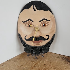 以下が含まれることがあります： 手描きの顔が描かれた楕円形の掛け時計。黒い髪、アーチ型の眉、茶色の目、目立つ黒い口ひげが特徴です。時計の下部には3つの黒いバラと葉の彫刻が施されており、金色の時計の針が付いています。