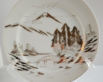 May include: A white ceramic plate with a gold and brown Japanese landscape design. The design features a mountain, trees, a bridge, and a river.