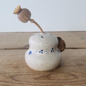 Puede incluir: Un jarrón de cerámica blanco con diseños florales azules y un acento de corcho. El jarrón tiene varios agujeros en la parte superior para sostener flores secas. Una cápsula de amapola seca está en el jarrón.