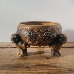 May include: A brown ceramic planter with a decorative floral pattern and three small figures supporting the base. The planter has a wide opening and is set on a wooden surface.