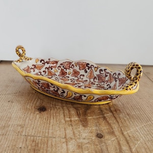 May include: Oval ceramic serving dish with a yellow rim and handles. The dish features a cream-colored base with a brown and red floral pattern. The handles have a yellow and brown spotted design.