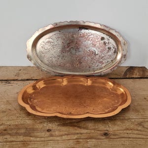May include: Two vintage metal trays. The top tray is silver-colored, oval, and features an ornate embossed design. The bottom tray is copper-colored and has a scalloped edge. Both trays are displayed on a wooden surface.