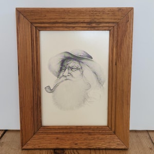 May include: A framed pencil sketch of an elderly man with a long white beard, hat, and pipe. The artwork is in a wooden frame, resting on a wooden surface. The man's face has detailed shading.