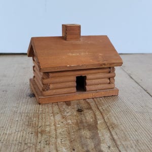 May include: A small, brown wooden log cabin model featuring a gabled roof, a central chimney, and a rectangular doorway opening. The structure is built with horizontal log-style pieces and sits on a flat, rectangular wooden base.