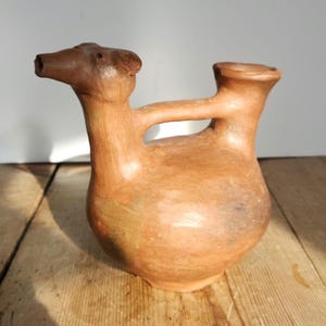 May include: A handcrafted, terracotta-colored pottery vessel with a unique design. The vessel features a rounded body, a spout, and a handle connecting to a cylindrical opening. The pottery is displayed on a wooden surface.