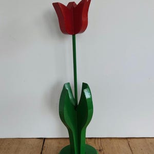 May include: A wooden tulip sculpture with a vibrant red flower head and a long green stem. The base and leaves are also green, creating a striking contrast against the wooden floor and white background. The tulip is a decorative item.