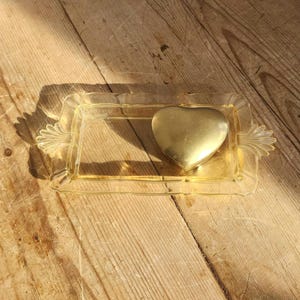 May include: A clear, rectangular, yellow-tinted tray with decorative handles, holding a gold heart-shaped box. The tray sits on a wooden surface, with sunlight casting shadows across the scene.