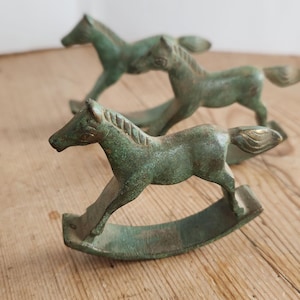 May include: Three small, weathered green metal rocking horse figurines. Each horse features a textured mane, a curved rocker base, and a distinct patina finish. The horses are arranged in a staggered formation, showcasing their identical design.