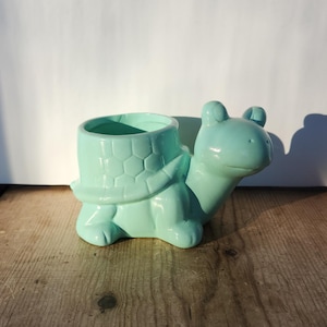 May include: A light teal ceramic turtle-shaped planter. The turtle has a textured shell, a smiling face, and a round opening for plants. The planter is on a wooden surface.
