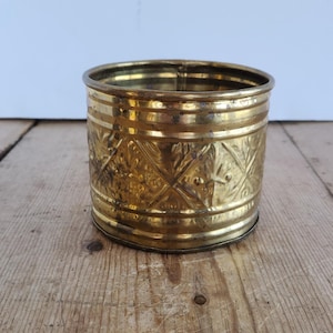 Puede incluir: Una maceta cilíndrica de latón con un patrón decorativo de diamantes en relieve alrededor del exterior. La superficie metálica muestra un acabado dorado pulido con sutiles signos de antigüedad y desgaste, incluyendo una ligera oxidación.