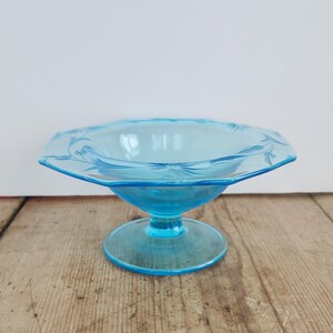 Puede incluir: Un cuenco de postre de cristal azul claro con base de pedestal. El cuenco tiene forma cuadrada con borde festoneado y diseño floral grabado. El plato se exhibe sobre una superficie de madera.