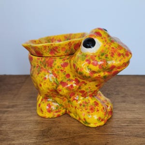 Peut inclure: Pot de fleurs en céramique en forme de grenouille, jaune avec des éclaboussures rouges et vertes. La grenouille a de grands yeux noirs et une large ouverture. Posé sur une surface en bois.
