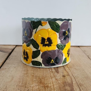 May include: A cylindrical metal planter featuring a cream-colored background decorated with hand-painted purple and yellow pansy flowers and green leaves. The top edge of the container has a scalloped design, and the exterior surface is smooth.