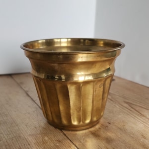May include: A gold-colored, fluted planter pot. The pot has a wide, rounded rim and a series of vertical grooves that run down the sides. The planter sits on a wooden surface.