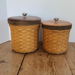 Peut inclure: Deux paniers cylindriques tressés avec des couvercles en bois brun clair et des boutons en métal argenté. Chaque panier présente une bande brun foncé sur le bord supérieur, fixée par de petits clous, et un corps en osier tressé naturel.